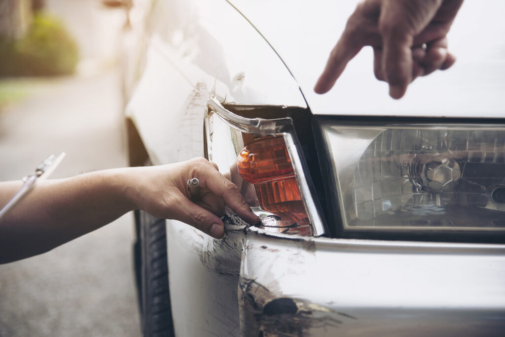A imagem mostra um close-up de uma parte da frente de um carro branco, focando em uma mão humana apontando para danos no veículo. A mão esquerda de outra pessoa está segurando a borda do para-choque dianteiro danificado perto do farol, onde a pintura está descascada e há sinais visíveis de um amassado. A luz indicadora de direção, que está integrada no para-choque, está rachada e danificada. O ambiente ao redor reflete um dia claro, possivelmente sugerindo uma cena externa com luz natural. Parece ser uma situação de avaliação de danos após um acidente de carro. Trata-se de um carro batido