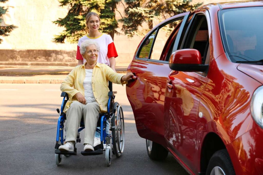 uma senhora idosa em uma cadeira de rodas ao lado de um carro vermelho, sendo auxiliada por uma mulher mais jovem, que parece ser sua cuidadora ou familiar.