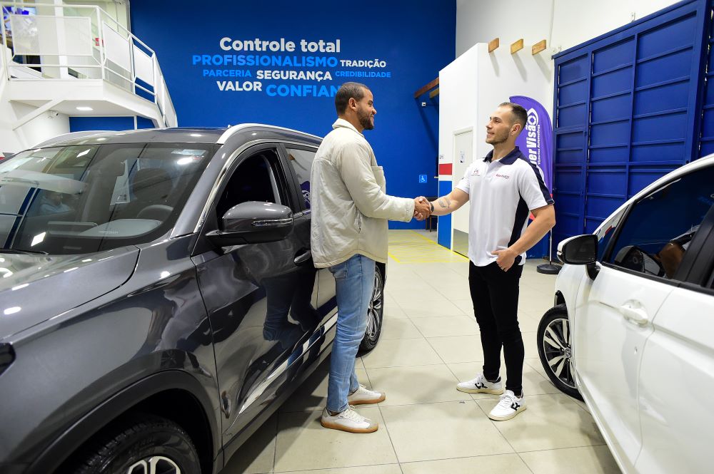 Cliente e consultor da Super Visão cumprimentando-se em unidade moderna de vistoria veicular, com veículos ao fundo e parede destacando valores como profissionalismo, segurança e confiança.