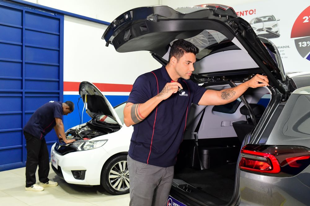 Vistoriadores uniformizados da franquia Super Visão realizando vistoria técnica em veículos com capô e porta-malas abertos em unidade moderna com identidade visual azul e vermelha.