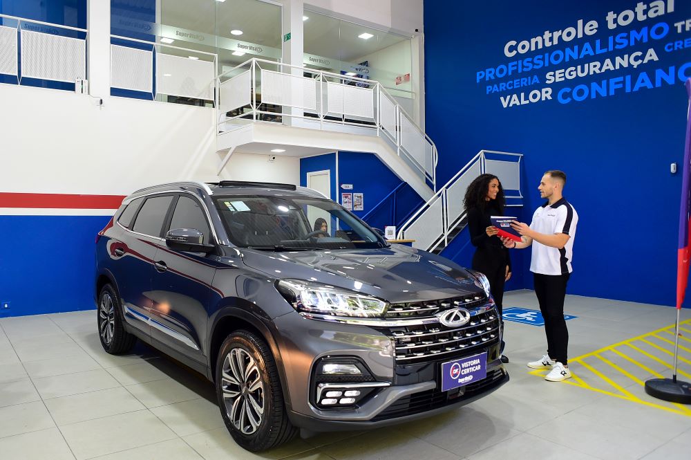 Atendimento personalizado em franquia Super Visão com cliente e consultor analisando documentação de vistoria veicular ao lado de SUV Chery cinza em unidade moderna com parede azul institucional.