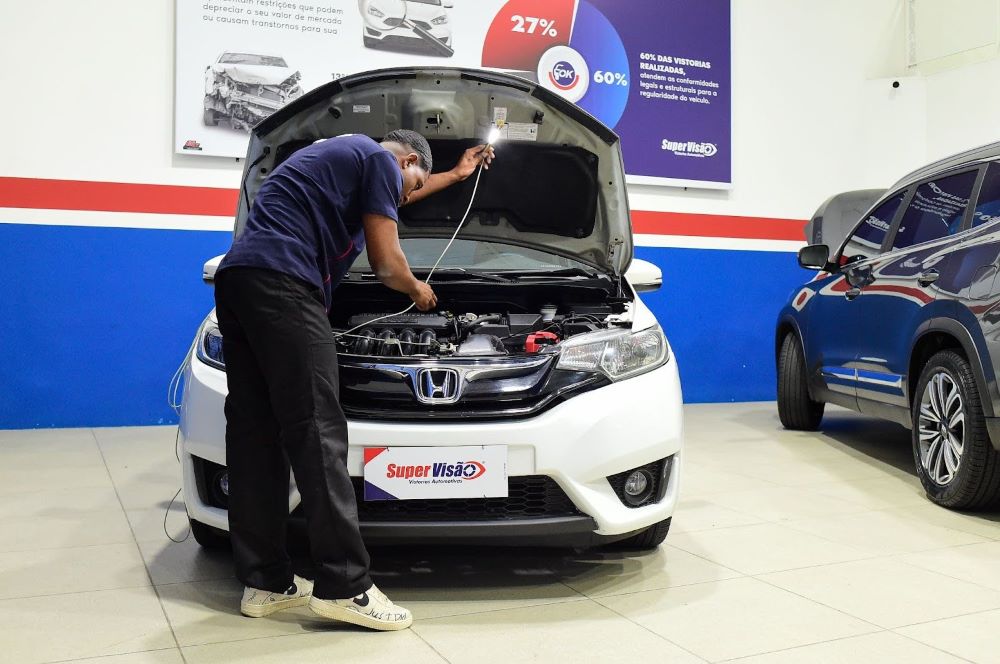 Vistoriador Super Visão, uniformizado com camiseta azul marinho, realiza inspeção no motor de carro em oficina, conforme regras da portaria 47/2025.