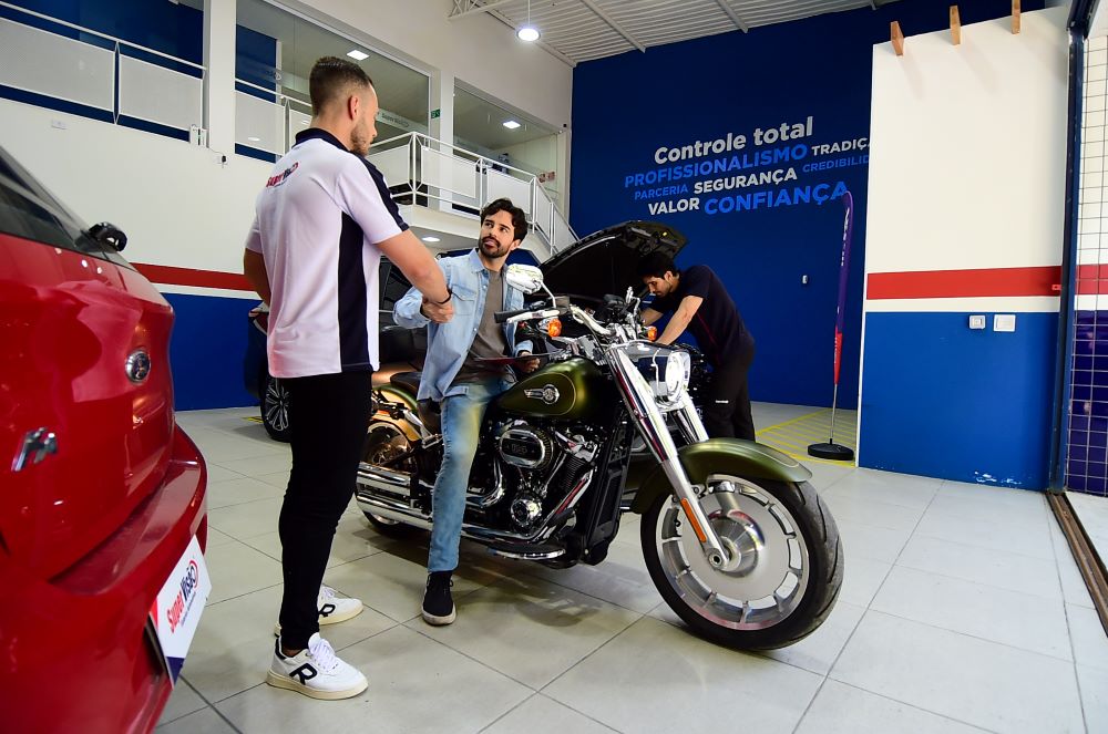 Interior de franquia Super Visão com técnicos uniformizados realizando vistoria simultânea em motocicleta Harley-Davidson e veículos. Ambiente profissional com painel informativo sobre controle total, profissionalismo, segurança e credibilidade em vistoria veicular.