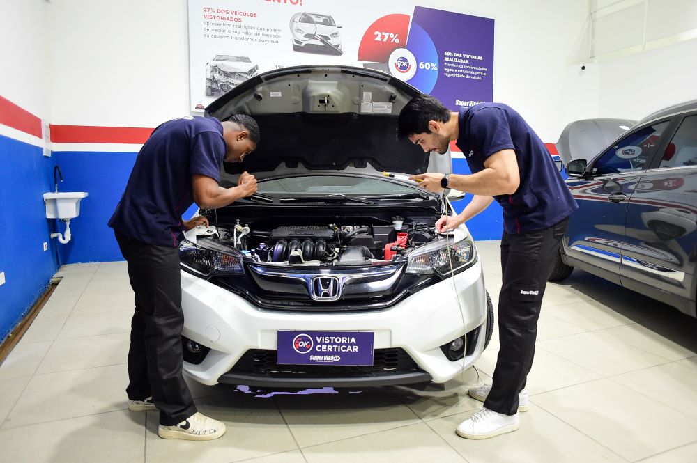 Técnicos da Super Visão realizando vistoria veicular simultânea em dois automóveis. No ambiente profissional com paredes azuis e vermelhas, uniformizados examinam capô aberto de Honda branco e porta-malas de SUV cinza. Painel informativo ao fundo exibe estatísticas de conformidade de vistorias. Demonstra profissionalismo, precisão e estrutura para franqueados crescerem.
