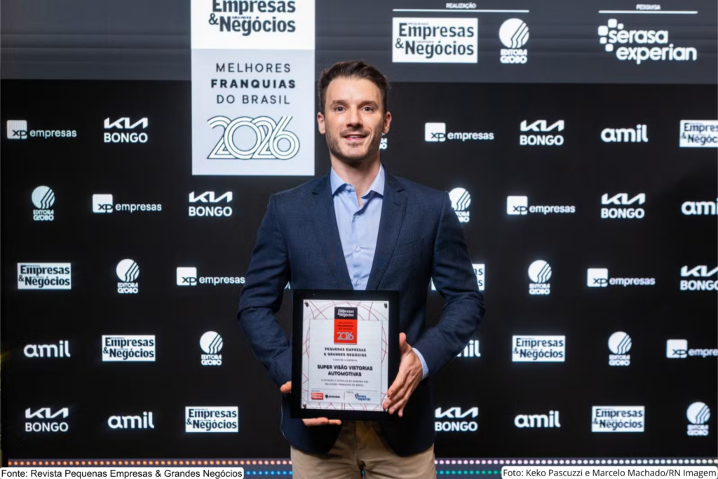 Representante da Super Visão segura placa da premiação Melhores Franquias do Brasil 2026, da Pequenas Empresas & Grandes Negócios, em frente ao painel oficial do evento.