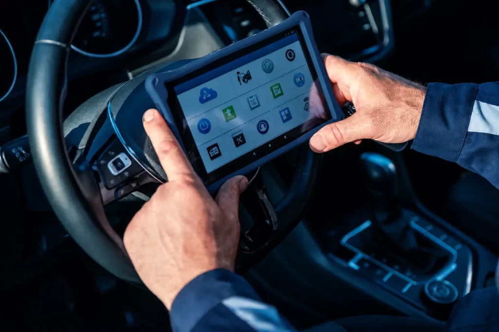 Mecânico com as mãos usando um tablet com tela de Diagnóstico Eletrônico Automotivo dentro do painel do carro, com ícones e opções de testes na interface.