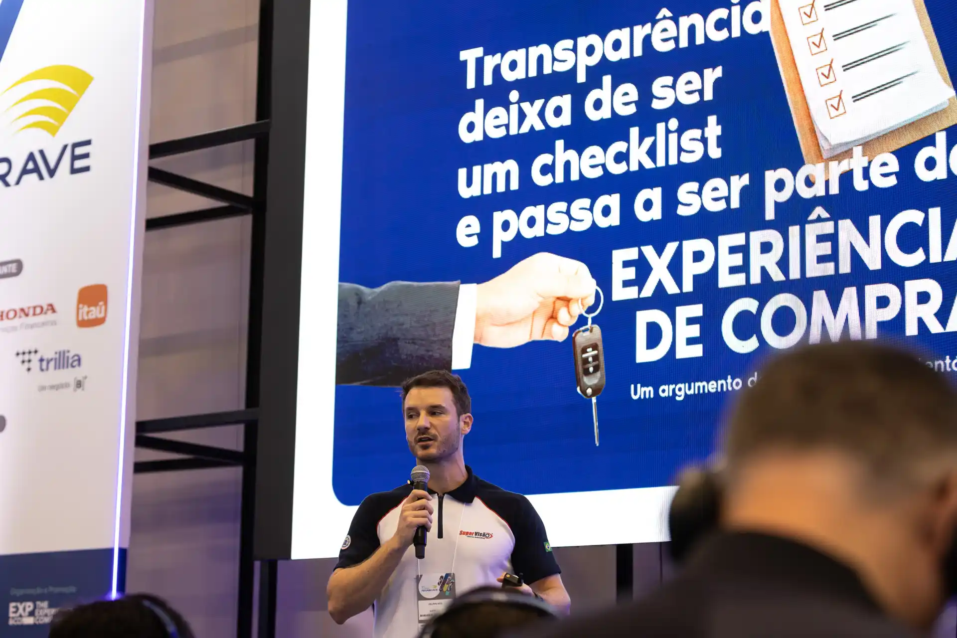 Fenabrave 2026: Felippe Reis, gestor comercial nacional da Super Visão Vistorias Automotivas, fala ao público em um evento com telão de apresentação e participante assistindo ao fundo.