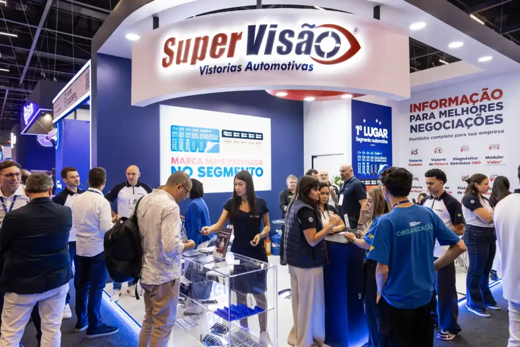 Participantes em um estande de feira automotiva, com placa “SuperVisão Vistorias Automotivas” e painéis de informações sobre serviços e resultados, em ambiente de evento com público circulando. Fenabrave 2026